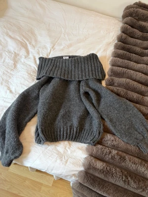 Grå off shoulder stickad tröja - Mysig mörkgråmelerad stickad tröja från Monki med bred off shoulder-krage och långa ärmar. Tröjan har ribbstickade muddar och är perfekt för en trendig och avslappnad look. Materialet är mjukt och värmande, perfekt för kyliga dagar.