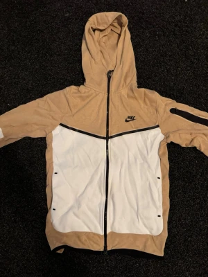 beige och vitt Nike Tech - Snygg nike tech  i beige och vitt med dragkedja framtill och svart Nike-logga på bröstet. Huva och långa ärmar, samt svarta detaljer och fickor framtill. Perfekt för en sportig och avslappnad stil.
