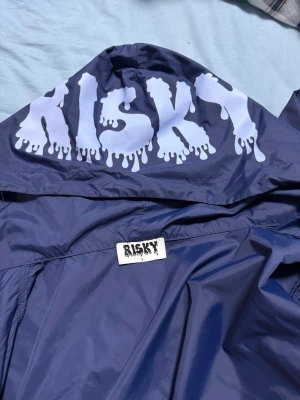 Blå vindjacka från Risky - Snygg blå vindjacka från Risky med vit logga i drip-style på ryggen och liten logga på ärmen. Jackan har dragkedja framtill och huva. Tillverkad i lätt och glansigt material, perfekt för blåsiga dagar.