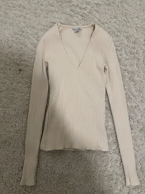 Beige ribbad v-ringad topp - Snygg beige ribbad topp från Aimsu med v-ringning och lång ärm. Toppen är figurnära och har en stilren design som passar perfekt till jeans eller kjol. Materialet är mjukt och stretchigt för en bekväm känsla.