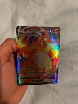 Charizard VMAX Pokémonkort - Säljer ett glansigt Charizard VMAX Pokémonkort med färgstark holografisk yta. Kortet har motiv av Charizard i orange och rött, med HP 330 och attacker som Claw Slash och G-Max Wildfire. Perfekt för samlare eller Pokémonfans. 