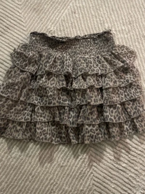 Leopardmönstrad volangkjol - Kort volangkjol med leopardmönster i bruna och beige nyanser. Kjolen har bred resår i midjan och flera lager volanger som ger en snygg och luftig känsla. Tillverkad i lätt syntetmaterial.