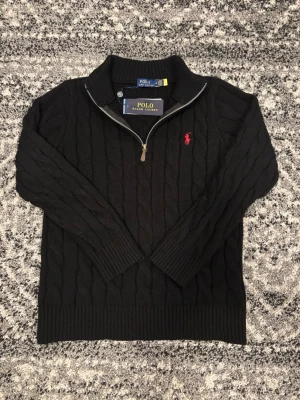 Svart kabelstickad tröja Polo Ralph Lauren - Svart kabelstickad tröja från Polo Ralph Lauren med halv dragkedja och röd broderad logga på bröstet. Tröjan har ribbade muddar och hög krage, perfekt för lager-på-lager. Klassisk och stilren design. Tröjan är i storlek M men passar även folk i stoleken S. Hör av er om ni har några frågor eller funderingar!