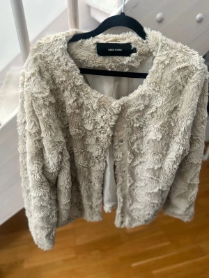 Beige teddyjacka från Vero Moda - Mysig beige teddyjacka från Vero Moda med rund halsringning och knappar framtill. Jackan har en fluffig, mjuk struktur och är perfekt för dig som vill ha en trendig och bekväm look. Passar till många olika outfits och ger en cool vibe.