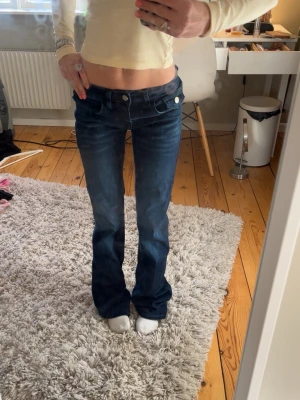 Mörkblå bootcut jeans - Säljer ett par mörkblå bootcut jeans med låg midja och snygga broderade detaljer på bakfickorna. Jeansen har vita knappar och klassisk femficksdesign. Perfekta för dig som gillar en retro vibe och vill sticka ut lite extra.