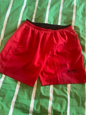 HUGO BOSS badbyxor XL - Snygga röda badbyxor från BOSS med svart resår i midjan och diskret BOSS-logga på vänster ben. Strl XL