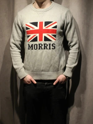 Grå Morris tröja - Grå stickad tröja från Morris med Union Jack-flagga och texten 'MORRIS' framtill. Tröjan har rund halsringning, ribbade muddar och är tillverkad i ett mjukt material. Perfekt för dig som gillar en stilren look 