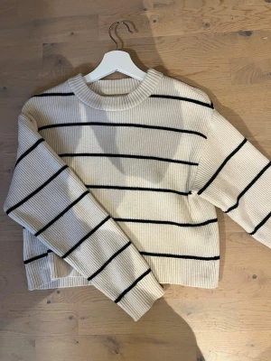 Randig stickad tröja från Zara - Snygg stickad tröja från Zara i beige med svarta ränder. Tröjan har rund halsringning, ribbstickade muddar och en avslappnad passform. Perfekt för en trendig och bekväm look.