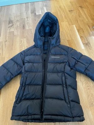 Svart dunjacka från Peak Performance XS - Säljer en svart dunjacka från Peak Performance med huva och dragkedja. Jackan har en puffer-design med breda kanaler och logga på bröstet. Xs i storlek och jackan är i ganska bra skick. Kedjan krånglar lite men den funkar om man gör det rätt. Säljer då den inte gått till användning.