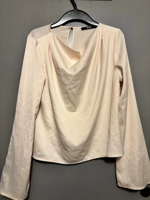 Ljusbeige draperad blus Bik Bok - Stilren ljusbeige blus från Bik Bok med draperad halsringning och långa ärmar. Tillverkad i ett mjukt, lätt tyg som ger ett elegant fall. Knäppning bak i nacken. Perfekt för att lyfta din outfit.