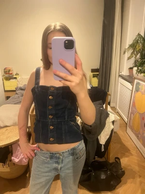 Mörkblå denim korsett med knappar - Säljer en mörkblå korsett-topp i denim med snygga träknappar framtill och breda axelband. Toppens passform är tight och går att shytstera längd. Storlek XS 