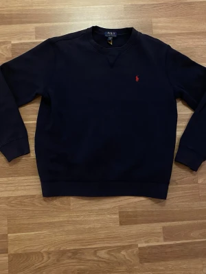Mörkblå sweatshirt från Polo Ralph Lauren - En stilren mörkblå sweatshirt från Polo Ralph Lauren med klassisk rund halsringning och röd broderad logga på bröstet. Tröjan har ribbade muddar vid ärmslut och nederkant. Perfekt för en avslappnad och snygg look.