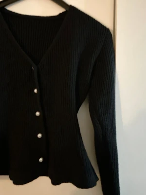 Svart ribbad tröja med pärlknappar - Snygg svart ribbstickad tröja med v-ringning och pärlknappar framtill. Koftan har långa ärmar och en figurnära passform. ALDRIG ANVÄND!