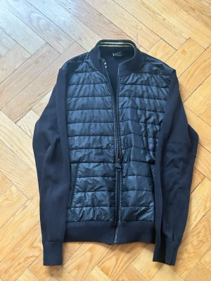 Svart cardigan med dragkedja BARBOUR - Snygg svart cardiganjacka med quiltad front och stickade ärmar samt rygg. Jackan har dragkedja framtill och ribbade muddar vid ärmslut och nederkant. Perfekt för lager-på-lager och en stilren look.