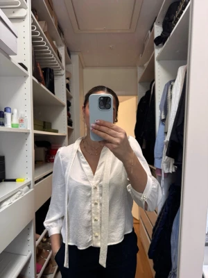 Blus Zara - Så fin vit blus med knytband i halsen och dekorativa pärlknappar framtill. Blusen har trekvartsärm och är tillverkad i ett lätt, strukturerat tyg som ger en elegant känsla. Perfekt för att skapa en chic look.