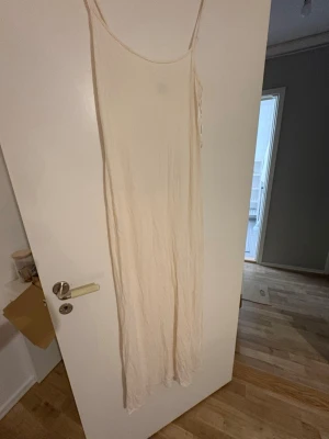 Lång ljusbeige klänning med smala band - En lång, ljusbeige klänning med smala axelband och fitted passform. Klänningen har en minimalistisk design och är tillverkad i ett tunt, lätt material som ger ett avslappnat intryck. Perfekt för varma dagar och enkel att styla med accessoarer. Den är stretchig och anpassar sig efter kroppen. Passar därför mindre och större storlek. 