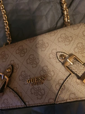 Beige axelremsväska från Guess - Snygg beige handväska från Guess med guldfärgade detaljer och kedjerem. Väskan har ett subtilt mönster med Guess-loggor över hela ytan och dekorativa spännen framtill. Perfekt för dig som vill ha en trendig accessoar med lyxig känsla.