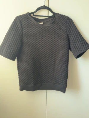 Svart strukturerad topp från H&M - Svart topp från H&M med korta ärmar och coolt strukturerat mönster i tyget. Rund halsringning och ribbade kanter vid hals och nederkant. Perfekt för dig som gillar en stilren men unik look.