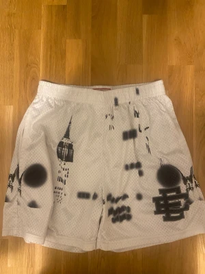 Vita meshshorts från Eric Emanuel med svart grafiskt tryck - Säljer ett par vita meshshorts med svart grafiskt tryck och spray paint-effekter. Shortsen har elastisk midja och är tillverkade i ett luftigt material som passar perfekt till sommaren. Unik streetwear-vibe med coola detaljer och logga på benet.