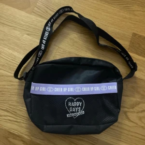 Svart axelväska Happy Days - Cool svart axelväska från Cheer Up Girl med justerbart band där det står 'CHEER UP GIRL'. Framsidan har ett broderat hjärta med texten 'HAPPY DAYS ENJOY&CO' och en lila detalj. Väskan är i nylon och har dragkedja upptill.
