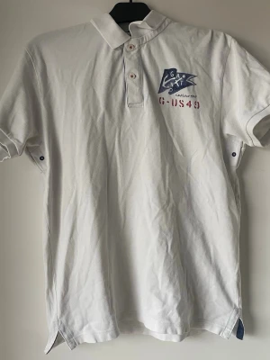 Vit pikétröja från Gant Storlek L - Klassisk vit pikétröja från Gant med blått och rött tryck på bröstet. Tröjan har korta ärmar, krage och knäppning med tre knappar framtill. Materialet är pikétyg i bomull som ger en sportig och fräsch look. Den lilla lappen som det står storleken på är lite riven men funkar endå 