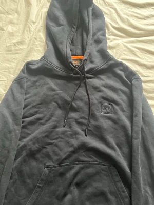 Svart hoodie från BOSS med logga - Snygg svart hoodie från BOSS med klassisk huva och dragsnören. På bröstet sitter en stilren svart patch med BOSS-loggan. Hoodien har en stor magficka och är gjord i mjukt bomullsmaterial som känns skönt mot huden. Perfekt för en avslappnad och trendig look.