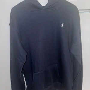 Mörkblå hoodie från Polo Ralph Lauren - Snygg mörkblå hoodie från Polo Ralph Lauren med klassisk huva och känguruficka. Liten vit broderad logga på bröstet. Tillverkad i mjukt bomullsmaterial som känns skönt mot huden. Perfekt för en avslappnad och stilren look. Saknar tags. De är avklippta. 
