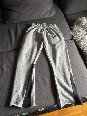Grå mjukisbyxor GALLEN DEPT. - Säljer ett par grå mjukisbyxor från GALLERY DEPT. med svart panel på insidan av benen och färgstänk som ger en unik streetwear-look. Byxorna har resår i midjan och tryckt logga på vänster sida. Perfekta för dig som gillar avslappnad och trendig stil.  (det står XL på storleken men dem är S egentligen)