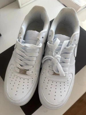Nike Air Force 1 vita sneakers - Klassiska Nike Air Force 1 sneakers i helvitt läder med perforerade detaljer på tån och sidorna. Skorna har en chunky sula, vita skosnören och den ikoniska swoosh-loggan på sidan. Perfekt för dig som gillar en clean och tidlös streetstil.