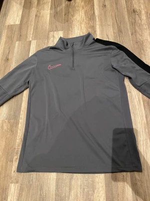 Grå Nike träningströja med dragkedja - Snygg grå långärmad träningströja från Nike med halv dragkedja framtill och svart detalj längs axlarna. Tillverkad i ett lätt och stretchigt funktionsmaterial som passar perfekt till träning eller uppvärmning. Diskret Nike-logga i rött på bröstet.