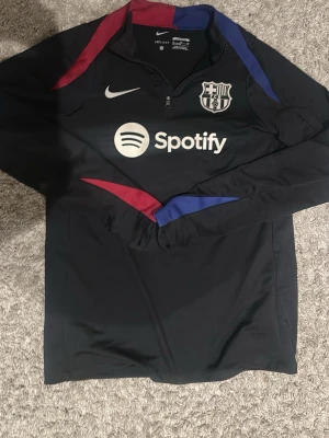 Nike FC Barcelona träningströja S - Säljer en svart långärmad träningströja från Nike med FC Barcelona-logga och Spotify-tryck på bröstet. Tröjan har färgdetaljer i rött och blått, halv dragkedja och är tillverkad i Dri-FIT polyester för bästa komfort vid träning.