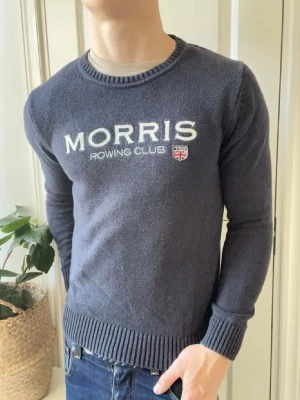 Mörkblå tröja från Morris Rowing Club - Mörkblå stickad tröja från Morris Rowing Club med vit broderad logga och liten brittisk flagga på bröstet. Rund halsringning och ribbade muddar vid ärmslut och nederkant. Perfekt för en sportig och stilren look.