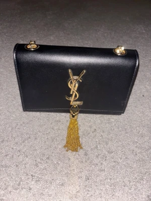 Svart YSL handväska med guldkedja - Snygg svart handväska från YSL i slätt läder med guldfärgad kedja och ikonisk YSL-logga framtill. Väskan har en dekorativ tofs i guld och stilren, rektangulär form. Perfekt för dig som gillar lyxiga accessoarer och vill sticka ut.  JAG ACCEPTERAR BUD!