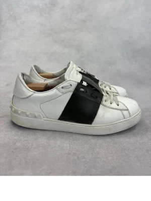 Vita och svarta sneakers från Valentino - Snygga vita sneakers från Valentino med en bred svart läderdetalj över sidan. Klassisk låg modell med vita snören och diskreta nitar på hälen. Perfekta för dig som gillar stilrena och exklusiva skor med en modern twist. Skorna är dock just nu lite smutsiga men en snabb rengöring så blir dom som på första bilden igen 