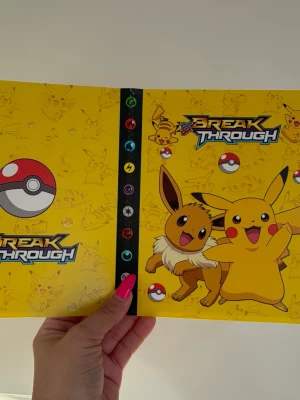 Pokémon Break Through samlarpärm - Gul Pokémon Break Through samlarpärm med motiv av Pikachu och Eevee på framsidan. Pärmen har plastfickor för att förvara och organisera samlarkort, perfekt för Pokémonkort. Tillverkad i plast och kartong med färgglada tryck och symboler från spelet. Plats för 240 st kort 