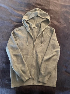 Grå ribbad hoodie - Säljer en stilren grå hoodie i ribbat material. Hoodien har dragkedja framtill, justerbar huva med snören och två fickor. Perfekt för en avslappnad look och enkel att matcha med andra plagg.