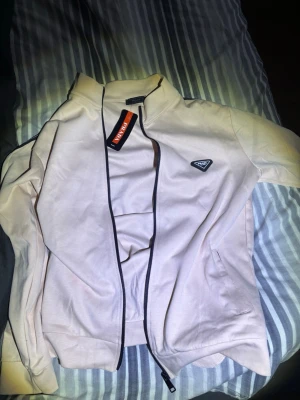 prada zip sweatshirt - Snygg ziptröja från Prada med svart kantband och ikonisk trekantslogga på bröstet. Tröjan har hög krage, dragkedja framtill och två sidofickor. Perfekt för dig som gillar exklusiv streetwear och stilren design. XL men passar M (liten i storlek)