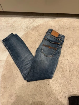 Blå jeans från Nudie Jeans - Säljer ett par klassiska blå jeans från Nudie Jeans med snygga slitningar och kontrastsömmar. Modellen har raka ben och fem fickor, samt Nudie-logga på bakfickan och läderpatch i midjan. Perfekta för en avslappnad och trendig look.