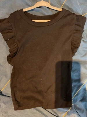 Svart topp med volangärmar - Svart topp med rund halsringning och snygga volangdetaljer på ärmarna. Toppens material känns mjukt och stretchigt, perfekt för en trendig och bekväm look. Passar till både jeans och kjol för en cool stil.