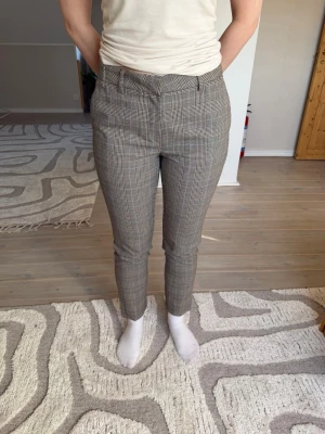 Rutiga kostymbyxor från Bondelid  - Snygga kostymbyxor med beige och svart rutmönste från Bondelid. Byxorna har smal passform, hällor för skärp och sidofickor. Perfekta för en stilren och trendig look. Materialet känns mjukt och följsamt.