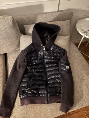 Svart Moncler cardiganjacka med huva - Snygg svart cardiganjacka från Moncler med quiltad, glansig front och ärmar i mjuk bomull. Jackan har huva, dragkedja framtill och två fickor med dragkedja. Moncler-logga på ärmen och ribbade muddar vid ärmslut och nederkant. Storlek M