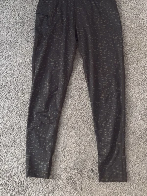 Svarta träningsbyxor från Newbody - Säljer ett par svarta träningsbyxor från Newbody med diskret camomönster. Byxorna har smal passform, elastisk midja och en ficka på sidan. Perfekta för gymmet eller löprundan.