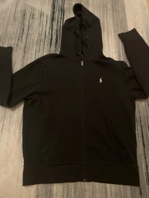 Svart hoodie från Polo Ralph Lauren - Klassisk svart hoodie med dragkedja från Polo Ralph Lauren. Har huva med dragsko, två fickor framtill och den ikoniska vita logotypen broderad på bröstet. Perfekt för en avslappnad och stilren look.