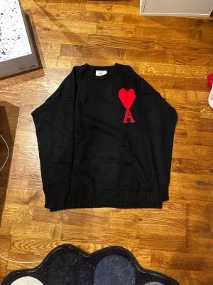 Svart stickad tröja från Ami Paris - Svart stickad tröja från Ami Paris med röd hjärtlogga och bokstaven A broderad på bröstet. Tröjan har rund halsringning och ribbade muddar vid ärmslut och nederkant. Perfekt för dig som gillar stilrena och trendiga plagg. Storlek M men skulle säga att den passar som L