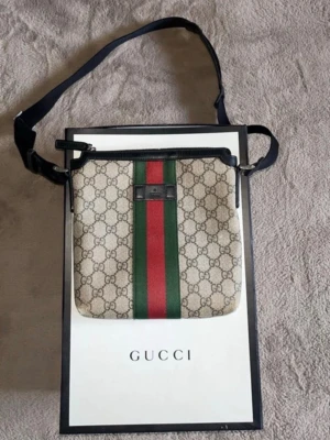 Gucci axelväska med GG-mönster - Snygg axelväska från Gucci i beige canvas med klassiskt GG-mönster och bred grön-röd rand i mitten. Väskan har svarta läderdetaljer och justerbar axelrem i svart textil. Perfekt för dig som vill ha en trendig och ikonisk accessoar.