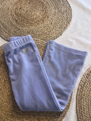 Ljuslila mjukisbyxor med hjärtdetalj - Säljer ett par ljuslila mjukisbyxor i mjukt velour tyg.  Byxorna har resår i midjan och en liten silverfärgad hjärtdetalj på ena sidan. Perfekta för chill dagar hemma eller när du vill vara extra bekväm. Storlek 104