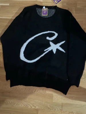 Svart stickad Corteiz tröja   - Svart stickad tröja från Corteiz med stor vit grafisk logga på framsidan och liten broderad detalj bak. Tröjan har rund halsringning och avslappnad passform. Perfekt för dig som gillar streetwear och vill sticka ut med en unik design.