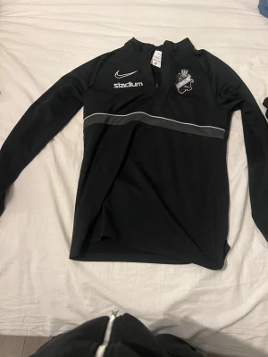 Svart Nike träningströja AIK - Svart långärmad träningströja från Nike med AIK-logga på bröstet och vita detaljer. Tröjan har halv dragkedja framtill, tryckta sponsorloggor från Stadium, Fabege och SVEA samt en vit vågig linje över bröstet. Perfekt för träning eller uppvärmning.