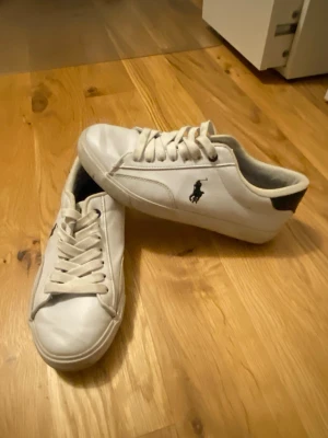 Vita sneakers från Polo Ralph Lauren - Snygga vita sneakers från Polo Ralph Lauren med klassisk låg profil, vita snören och svart logga på sidan. Skorna har en vit sula med Polo-tryck undertill och svart detalj vid hälen. Perfekta för en clean och stilren look.