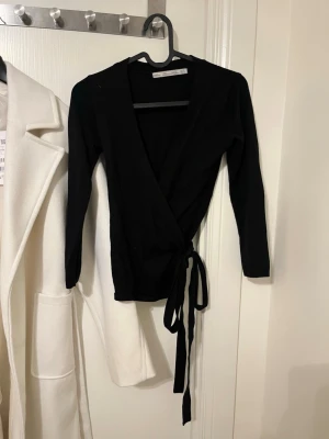 Svart omlottkofta med knytband - Svart omlottkofta med långa ärmar och knytband i midjan. Snygg och enkel design som passar perfekt till jeans eller kjol. Koftan har en figurnära passform och är tillverkad i ett mjukt, stretchigt material.